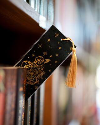 Decorative black bookmark with gold tassel and intricate design on a bookshelf.