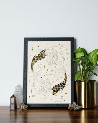 A framed artwork featuring a stylized illustration of a Pisces mermaid girls with flowing lines and oceanic details, displayed on a wooden table with a green potted plant and decorative items in the background.