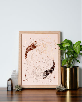 A framed artwork featuring a stylized illustration of a Pisces mermaid girls with flowing lines and oceanic details, displayed on a wooden table with a green potted plant and decorative items in the background.