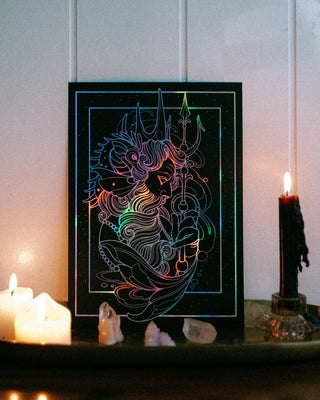 A black art print or panel with hologprahic silver foil of Poseidon, displayed on a dark tray with lit candles and crystals against a pale paneled wall.