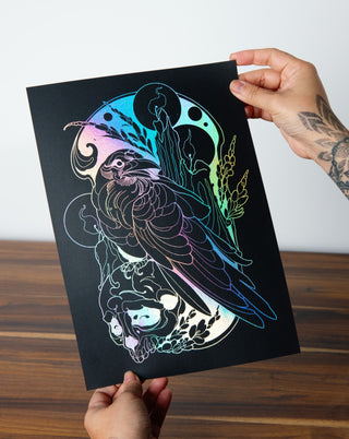 A tattooed hand holding up an art print in silver foil with a bird and skull on it.