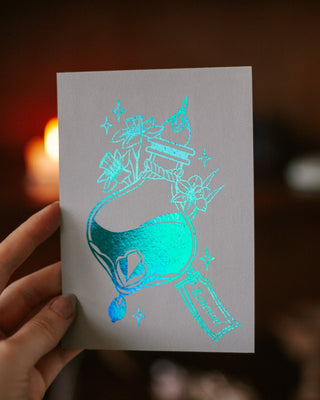 A hand holds a mini art printfeaturing a holographic, iridescent turquoise potion bottle with flowers. The background is a dark, warm, blurred environment with a glowing candle.