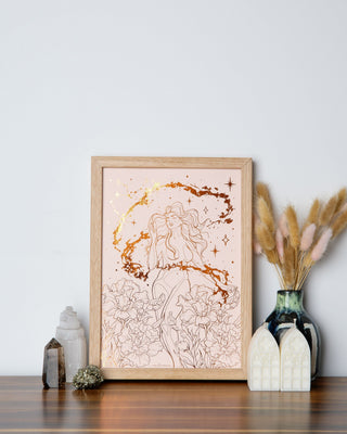 A framed art print with a Sagittarius woman, featuring a warm, orange foil accent, displayed on a wooden surface with decorative items around it.