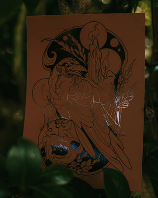 A brown paper print featuring an art illustration of a raven and candle decorative motifs, photographed outdoors among dark green leaves and branches.