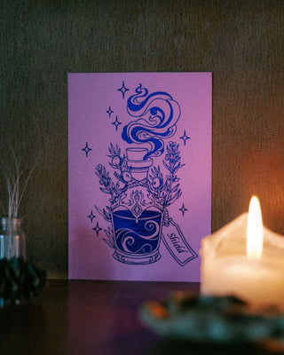A art print featuring a indigo foil illustration of a potion bottle with steam, herbs, and a tag reading 'Shield' on a purple background, displayed on a dark surface next to a lit candle.