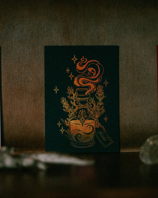 A dark holographic gold art print featuring a golden potion bottle with steam, surrounded by coffee plants and stars, displayed on a dark wooden surface.