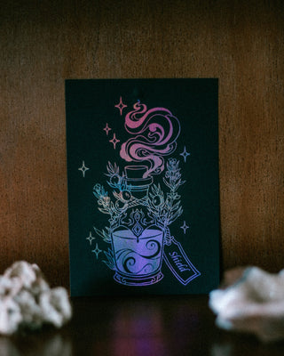 A black rectangular mini art print featuring a stylized glowing potion bottle illustration with purple iridescent accents, displayed upright on a dark wooden surface with blurred white crystals in the foreground.