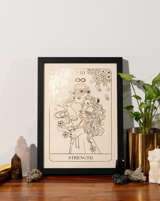 A framed tarot card art print featuring 'Strength' with gold foil line drawing on an off-white background, styled on a wooden surface next to a potted plant, crystals, and a small white figurine.
