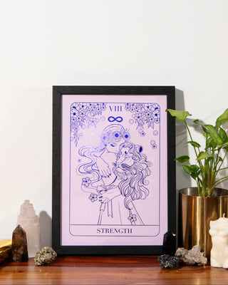 A framed tarot card art print featuring the 'Strength' card in purple line art on a light lavender background, styled on a wooden surface next to a potted plant and crystals.