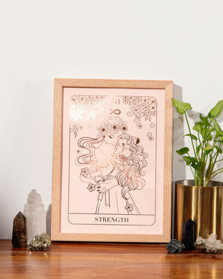 A framed tarot card print featuring 'Strength' with an illustration of a women embracing a lion, styled on a wooden surface against a white wall with crystals and a potted plant.