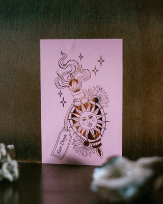 A mini A6 art print featuring a stylized sun, moon, stars, and a potion bottle illustration, displayed on a dark wooden surface with blurred decorative objects in the foreground.