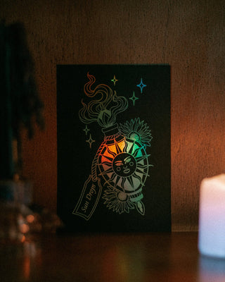 A black rectangular mini art print featuring a stylized sun illustration with a holographic foil effect, displayed on a dark wooden surface next to a lit white candle.