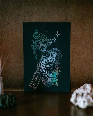 A dark teal art print featuring a stylized bottle labeled 'Sun Drops' with a sun face, floral details, and glowing star-like accents, displayed on a dark wooden surface with decorative crystals in the foreground.