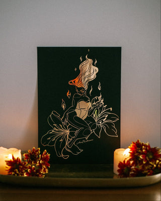 A black art print featuring gold foil drawing of a woman with flowing hair and flowers, styled on a tray with lit candles and red flowers against a neutral wall.