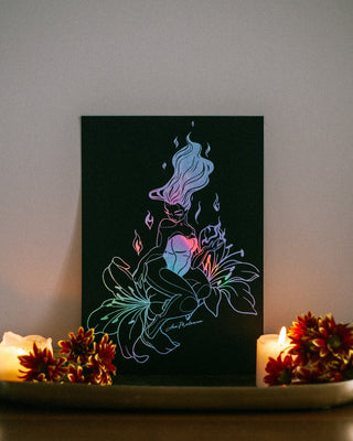 A black canvas art print featuring a glowing neon-style illustration of a woman with flowing hair and flowers, displayed on a shelf between lit candles and orange flowers against a neutral wall.