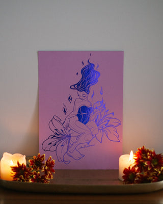 A vertical art print featuring indigo foil drawing of a woman seated on a flower, displayed on a shelf between two lit candles and small floral arrangements against a plain wall.