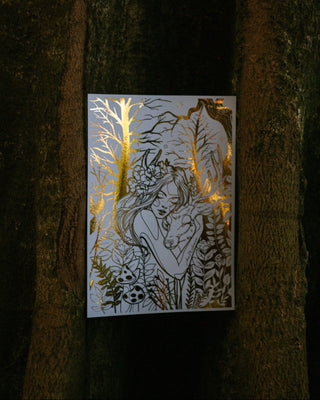 An art print featuring gold foil of a woman  hugging a deer in a forest, highlighted with shimmering gold foil accents, displayed against a dark, textured tree trunk background.