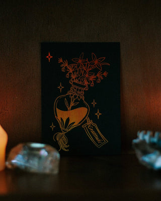 A black art print featuring a gold illustration of a heart-shaped bottle with flowers and a label reading 'Gratitude', displayed on a dark shelf with warm ambient lighting and crystals in the foreground.