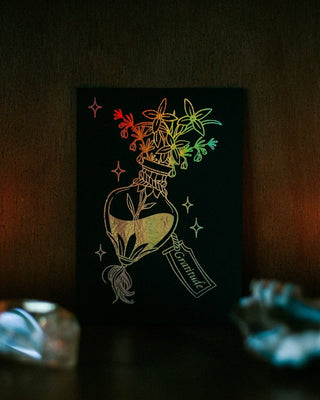 A black mini A6 art print or card featuring a potion bottle with flowers and a rainbow holographic foil effect, displayed on a dark shelf with candles and crystals around it.