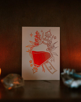 An A6 art print featuring a potion bottle and botanical wattle illustration ion a dark wooden surface with warm candlelight and blurred decorative objects in the foreground.