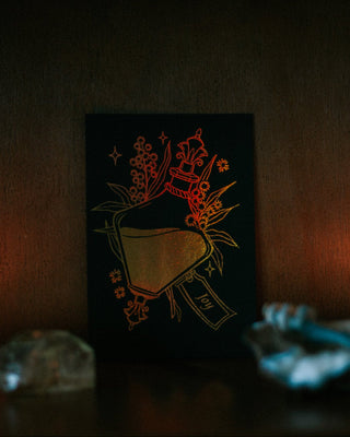 A small black art print featuring a potion bottle illustration in warm gold and red tones, displayed on a dark shelf with candles and crystals around it.