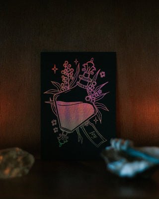 A black foil art print or card featuring a potion bottle with flowers, displayed on a dark shelf with candles and crystals around it.