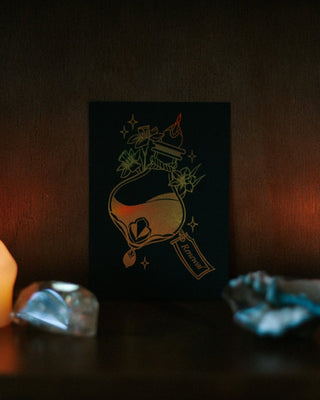 A small black art print or card featuring an illustration of a potion bottle and flowers, displayed on a dark shelf between lit candles and crystals.