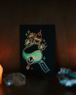 A black rectangular art print featuring a silver foil illustration of a potion bottle with small gold stars and florals, displayed on a dark wooden surface with warm ambient light and blurred candles around it.