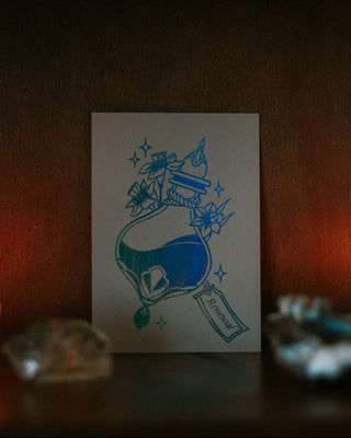 A A6 art print featuring an illustration of a blue potion bottle with the word 'Renewal' on a tag, displayed on a dark wooden shelf with warm ambient lighting and blurred decorative objects in the foreground.