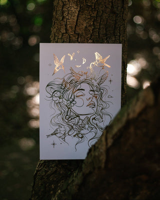 An art print featuring gold foil art illustration of a woman's face with birds, moons, and stars, displayed against a dark, blurred forest tree background.