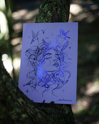 An art print featuring indigo foil illustration of a woman's face with birds and floral elements, printed on white paper and displayed against a tree trunk in a forest setting.
