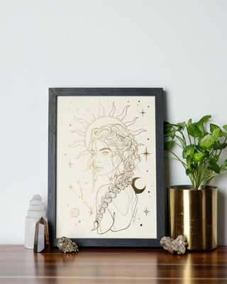 A framed print with an illustration of a female figure with celestial elements, placed on a wooden surface, with decorative items and a plant to the sides.
