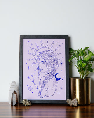 A framed print with an illustration of a Taurus girl with celestial elements, placed on a wooden surface, with decorative items and a plant to the sides.