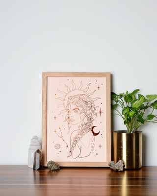 A framed print with an illustration of a female figure with celestial elements, placed on a wooden surface, with decorative items and a plant to the sides.