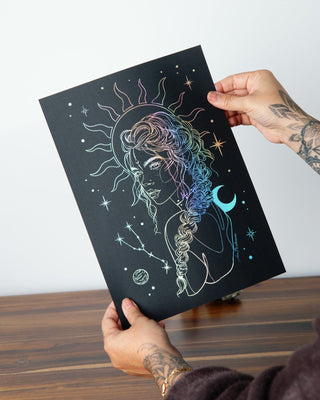 A person holds up an art print featuring a Taurus girl with stars, a sun, and a crescent moon, with holographic silver foil highlights. 