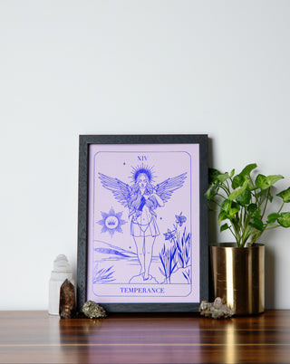 A framed print titled 'Temperance' depicting a being with wings pouring water from one vessel to another, set against a purple background, placed on a wooden surface with a plant and decorative objects around it.