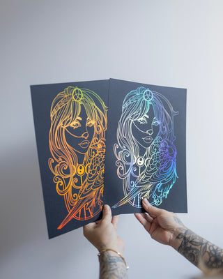 Two hands with tattoos hold up two Morrigan prints featuring holographic gold and silver foil set against a plain light gray background.