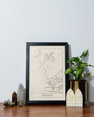 Framed artwork of 'The Fool' tarot card on a wooden surface with decorative items.