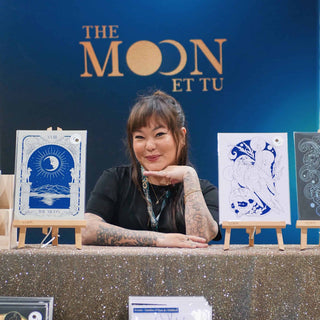 Person at a booth with 'The Moon Et Tu' branding, displaying art prints.