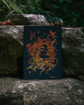 A dark  art print or card featuring an intricate line drawing of a woman's face with flowing hair and birds, placed on large rocks in a forest setting.