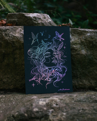 A dark art print featuring a profile illustration of a woman's face composed of flowing hair and surrounded by butterflies and birds, printed in iridescent pastel lines on a dark background. The print is propped up against large natural rocks with a blurred forest background.