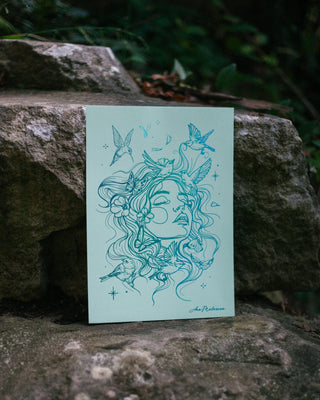 An art print featuring holographic foil drawing of a woman surrounded by birds and celestial motifs, displayed on a large stone outdoors with dark green foliage in the background.