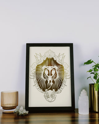 A framed art print featuring a detailed gold sting ray design on gold foil background, styled on a wooden table next to a potted plant, a candle, and a crystal cluster against a plain white wall.